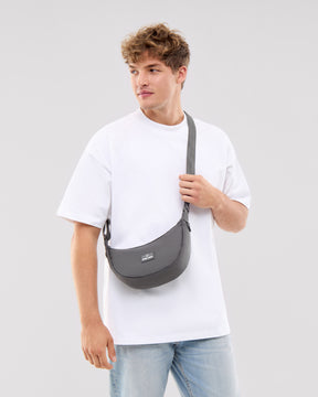 Mann trägt Half Moon Bag 'Ally Small' in dunklem Grau über der Schulter, kombiniert mit weißem T-Shirt und hellblauen Jeans