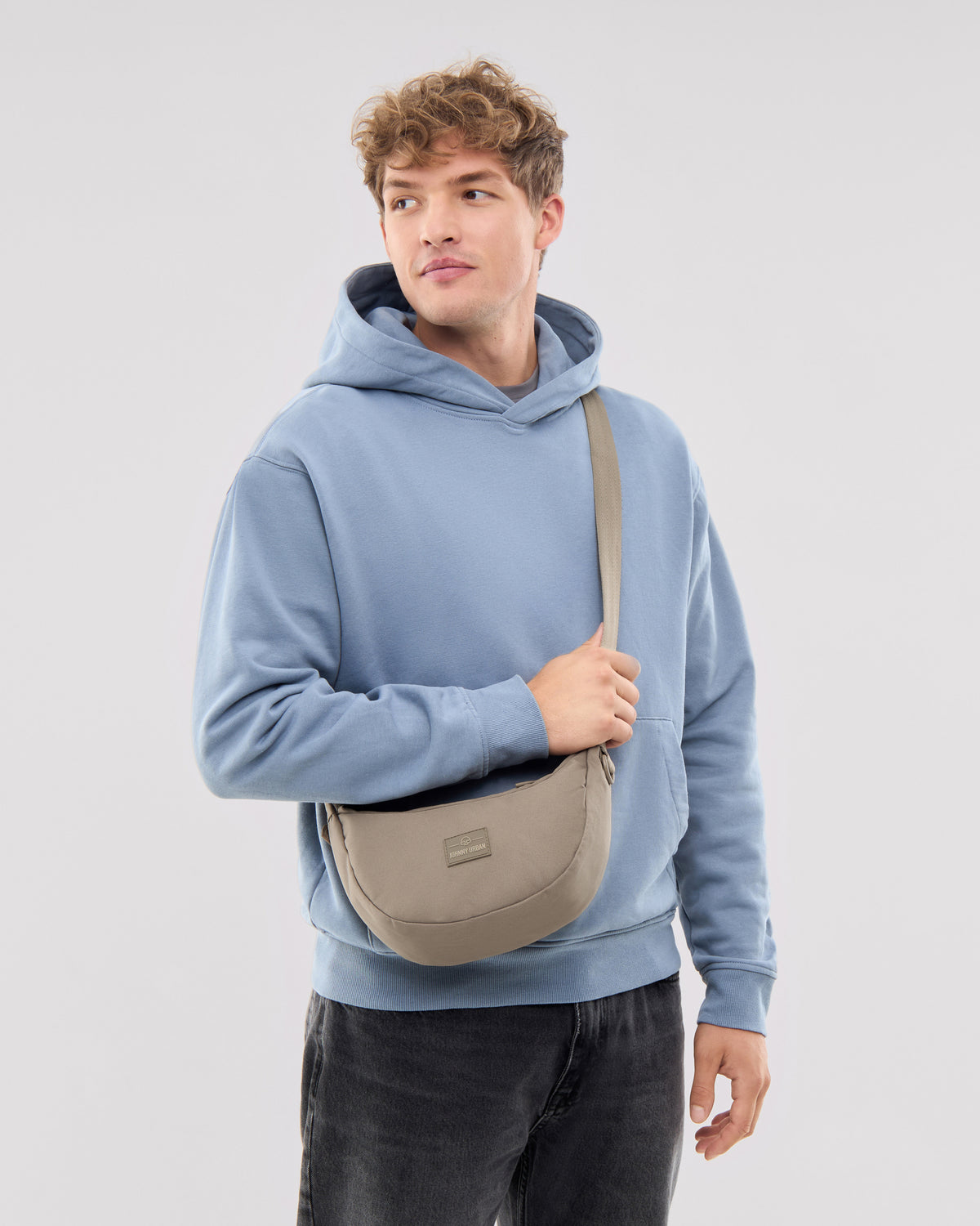 Mann trägt kleine Half Moon Bag 'Ally Small' in desert-grau über der Schulter, blaue Kapuzenjacke, schwarze Jeans