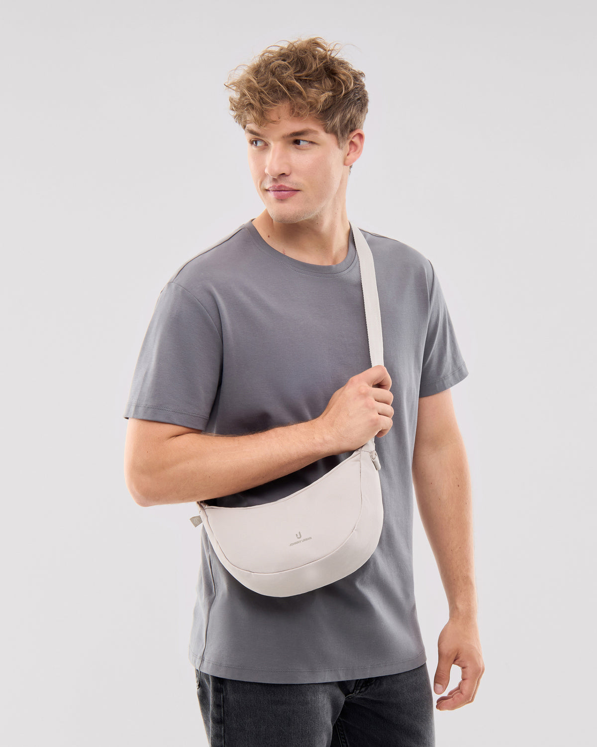 Mann trägt Johnny Urban Half Moon Bag 'Ally Small' in Sandfarben über die Schulter, grau T-Shirt, schaut seitlich