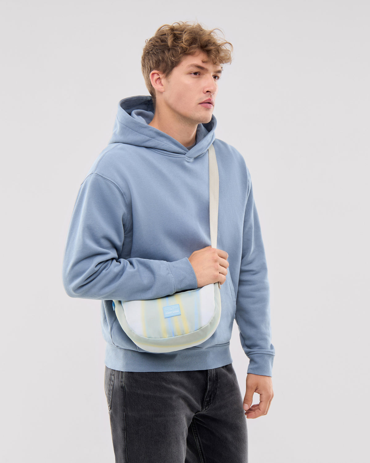 Mann trägt kleine Half Moon Bag 'Ally Small' in Sand mit Streifen und hellblauen Hoodie