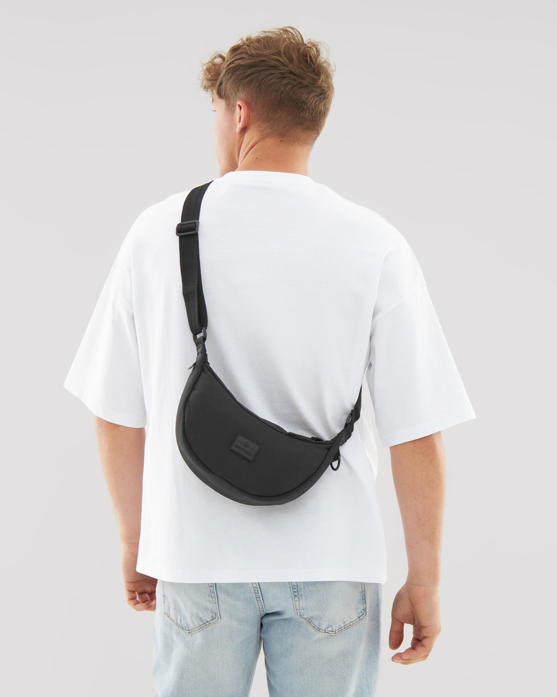 Mann trägt schwarze Half Moon Bag 'Kaia Small' quer am Rücken über weißem T-Shirt und blauer Jeans
