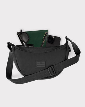 Half Moon Bag 'Kaia Small' von Johnny Urban, schwarze Halbmondtasche mit verstellbarem Schultergurt, geöffnet mit Brille, grünem Notizbuch, Stift und schwarzem Smartphone