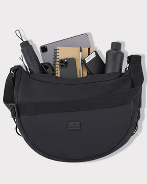 Schwarze Half Moon Bag 'Kaia XL' mit Schultergurt gefüllt mit Tablet, Handy, Notizbuch, Stift, schwarzer Geldbörse, Wasserflasche, Regenschirm und Sonnenbrille