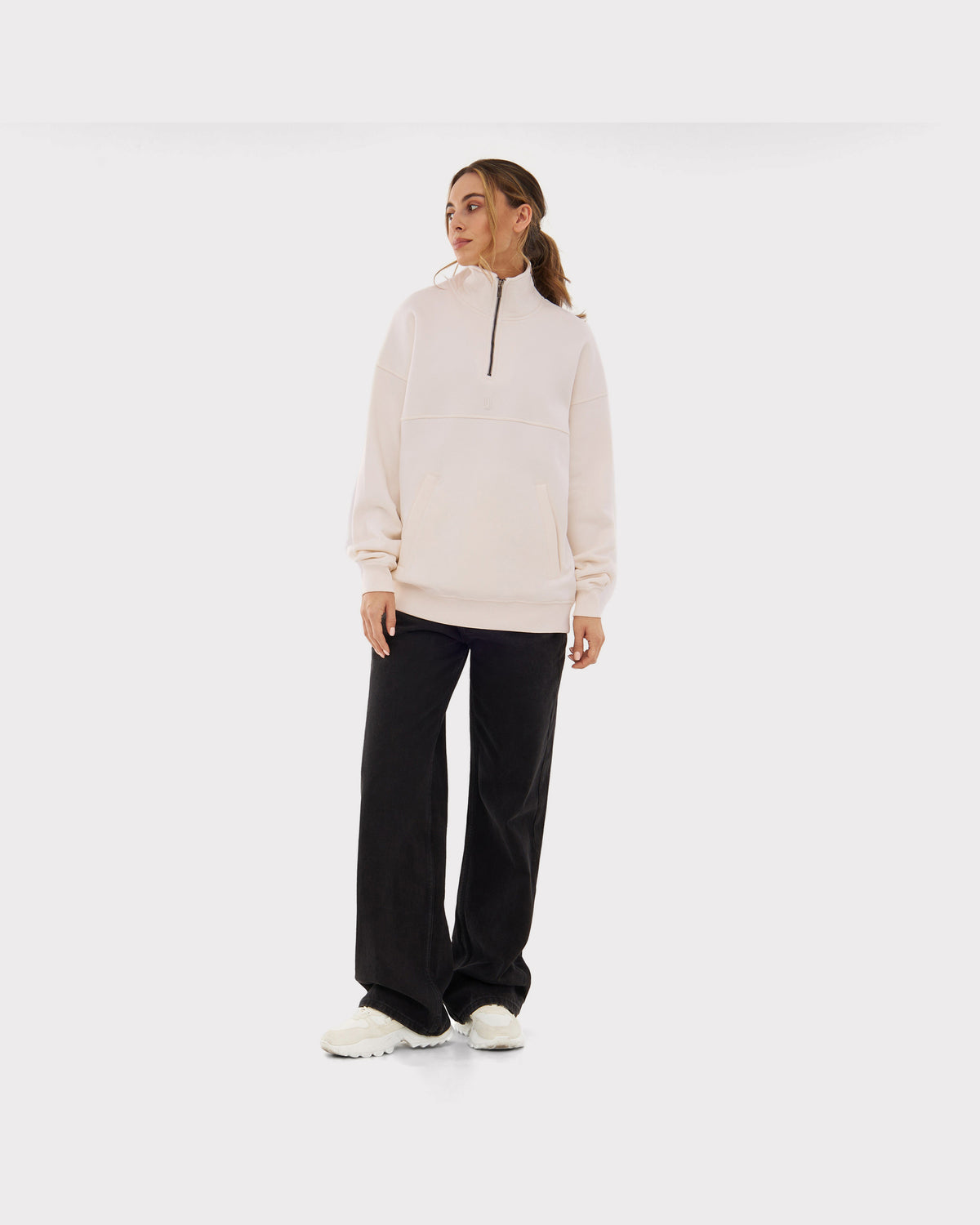 Frau trägt cremeweißen Half Zip Sweatshirt 'Cooper' mit schwarzem Reißverschluss, kombiniert mit schwarzen weiten Hosen und weißen Schuhen