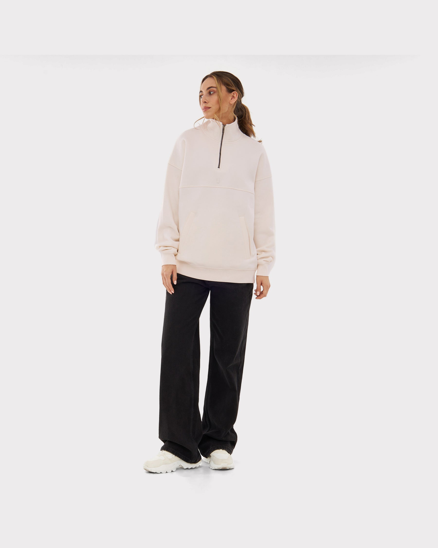 Frau trägt cremeweißen Half Zip Sweatshirt 'Cooper' mit schwarzem Reißverschluss, kombiniert mit schwarzen weiten Hosen und weißen Schuhen