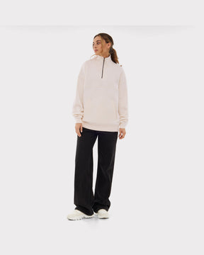 Frau trägt cremeweißen Half Zip Sweatshirt 'Cooper' mit schwarzem Reißverschluss, kombiniert mit schwarzen weiten Hosen und weißen Schuhen