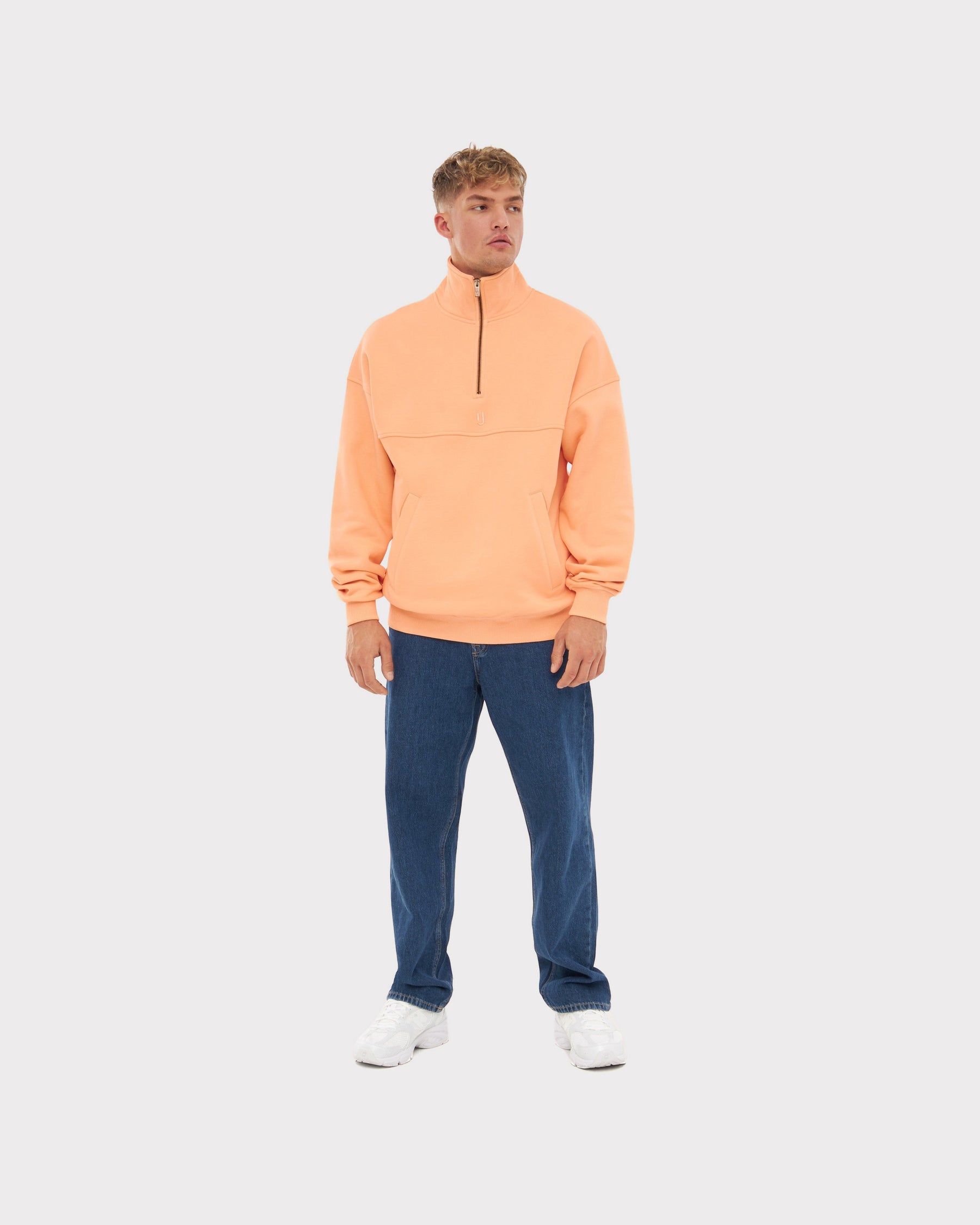 Mann trägt Johnny Urban Half Zip Sweatshirt 'Cooper' in Pfirsichfarbe, kombiniert mit blauer Jeans und weißen Schuhen