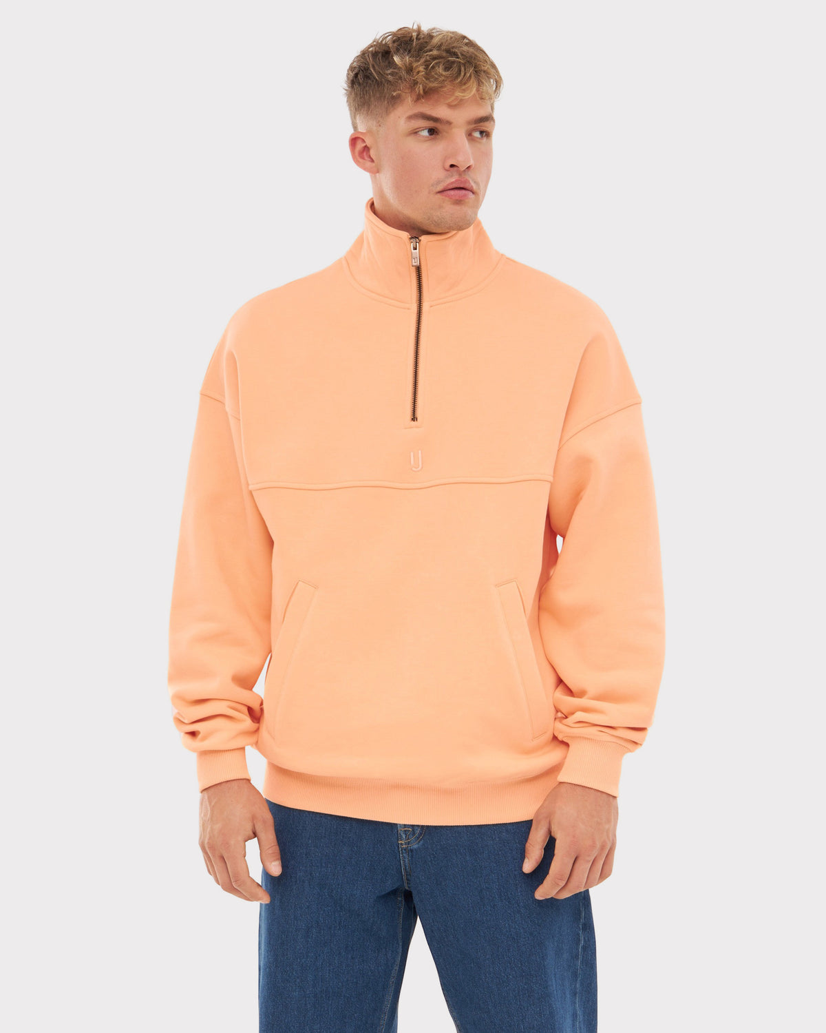 Mann trägt pfirsichfarbenen Halb-Zip-Sweatshirt 'Cooper' mit Stehkragen und Reißverschluss, kombiniert mit blauer Jeans