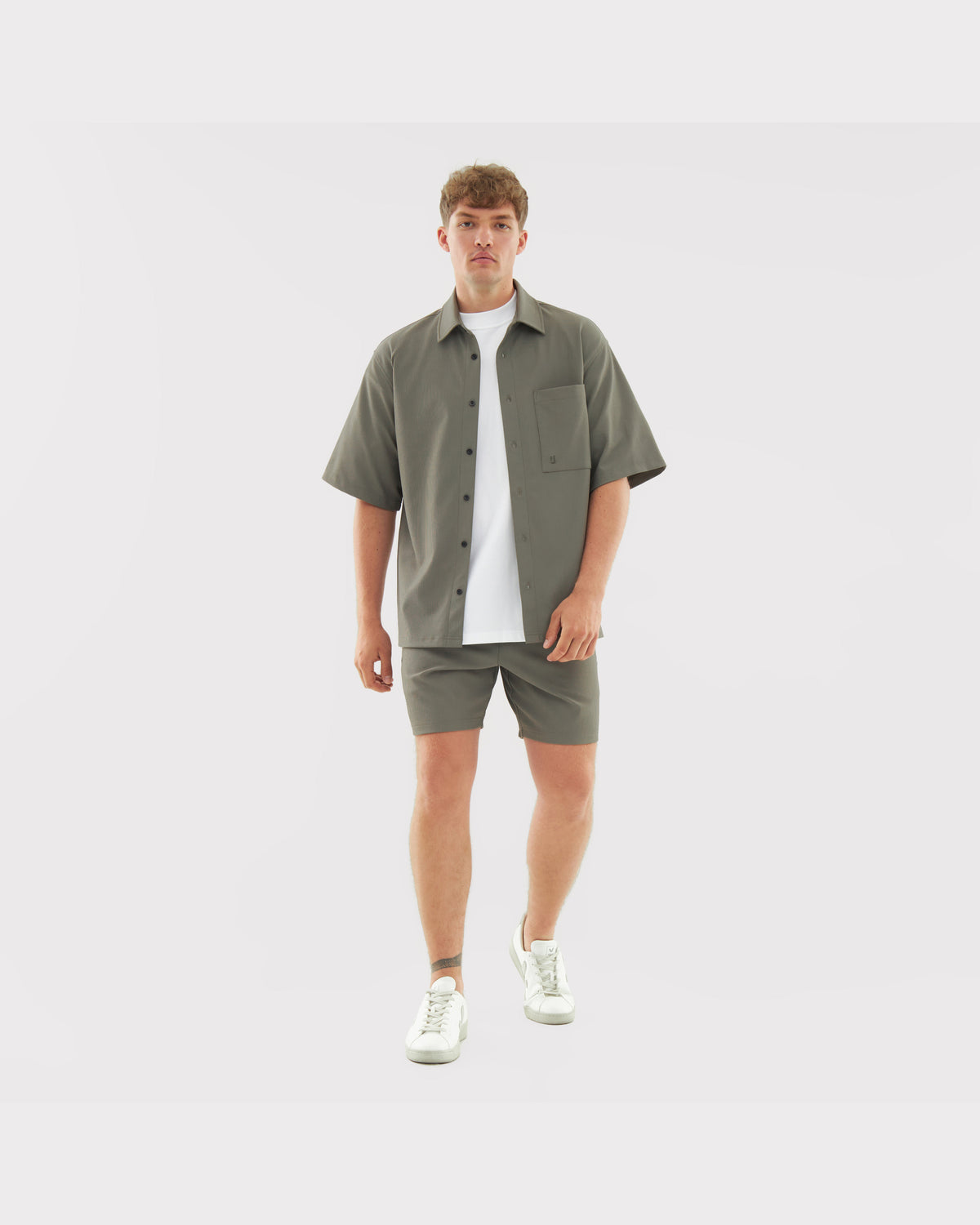 Männliches Model trägt lockeres, olivgrünes Hemd und passende Shorts aus dem Set 'Mason' mit weißem T-Shirt und weißen Sneakern