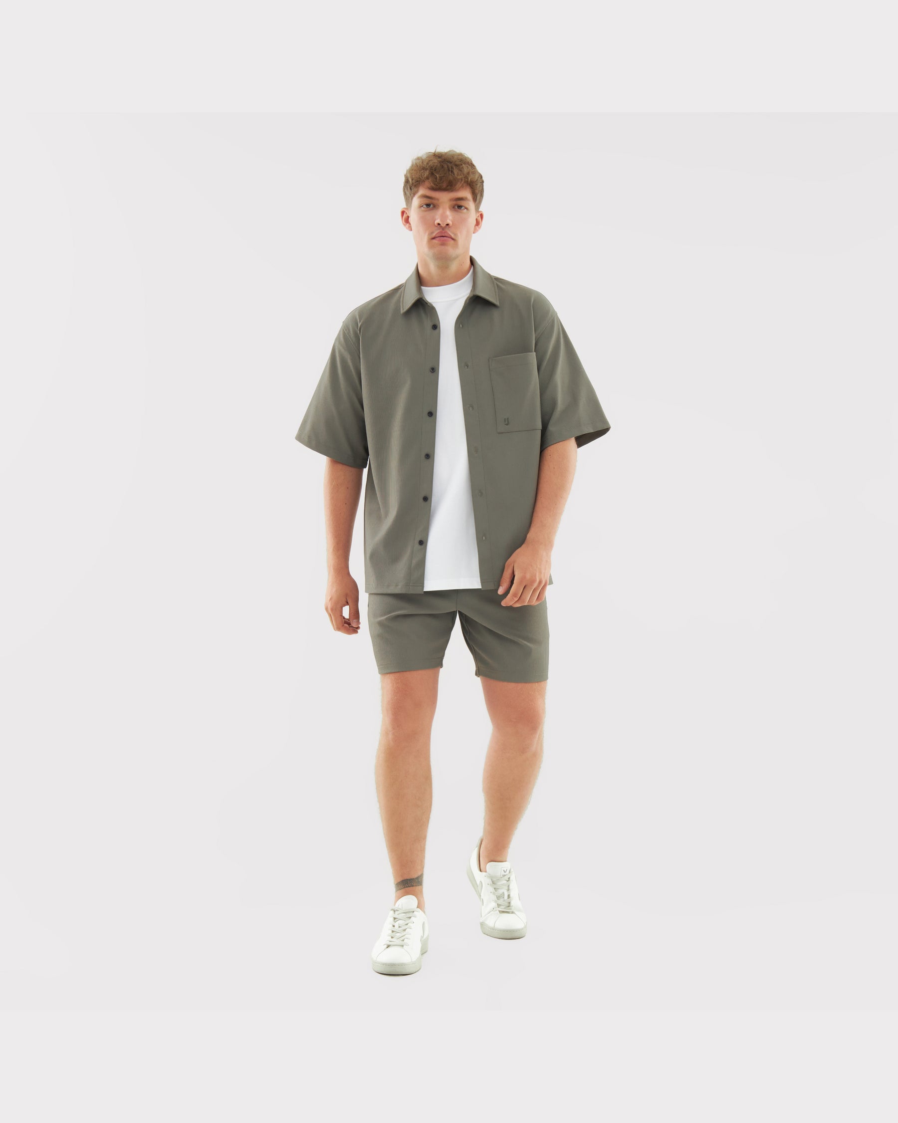 Männliches Model trägt lockeres, olivgrünes Hemd und passende Shorts aus dem Set 'Mason' mit weißem T-Shirt und weißen Sneakern