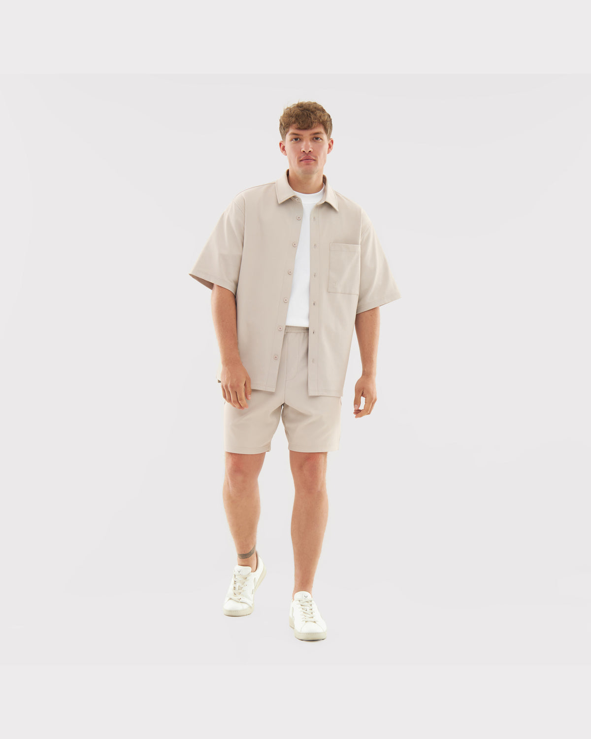 Männliches Modell trägt sandfarbenes Hemd 'Mason' mit kurzen Ärmeln und umgeschlagener Knopfleiste, kombiniert mit passenden Shorts und weißen Sneakern