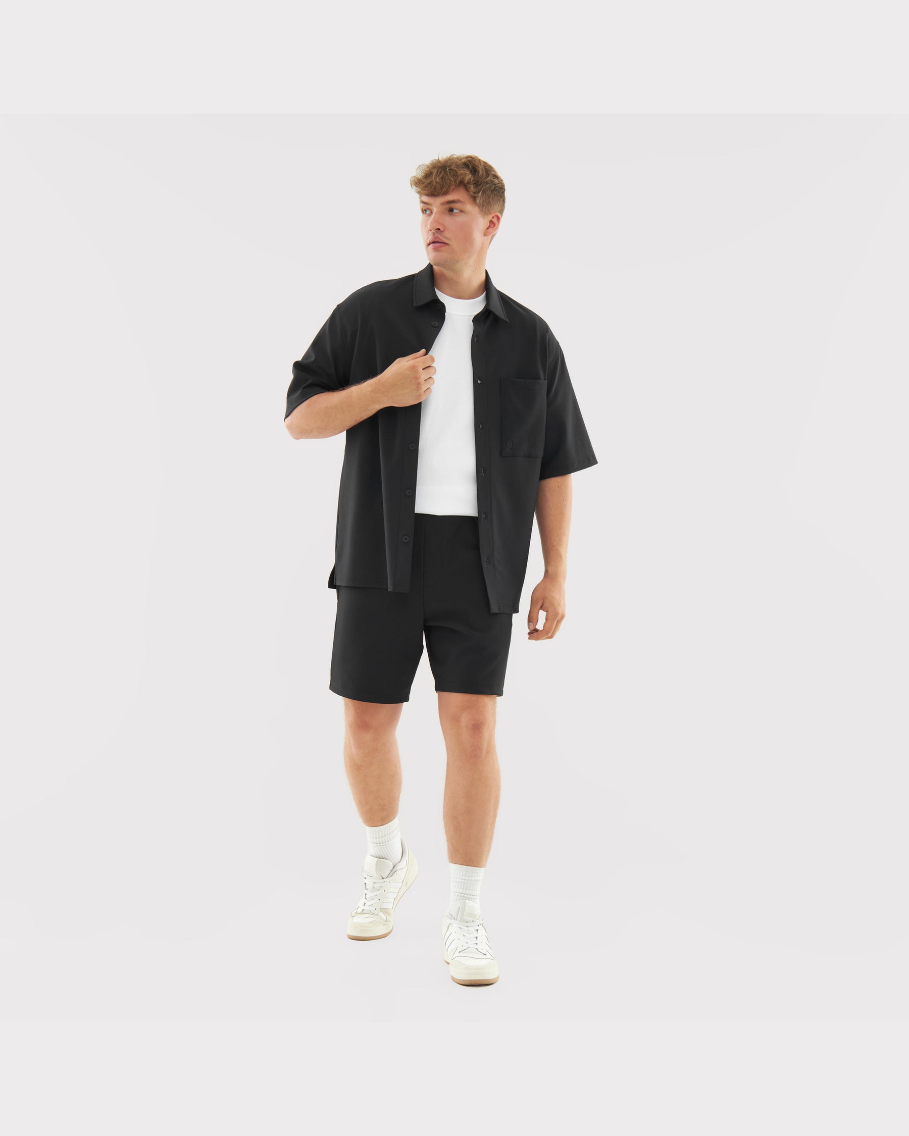 Mann trägt schwarzes, locker sitzendes Hemd und Shorts aus dem Set 'Mason', kombiniert mit weißem T-Shirt und Sportschuhen