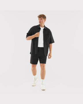 Mann trägt schwarzes, locker sitzendes Hemd und Shorts aus dem Set 'Mason', kombiniert mit weißem T-Shirt und Sportschuhen
