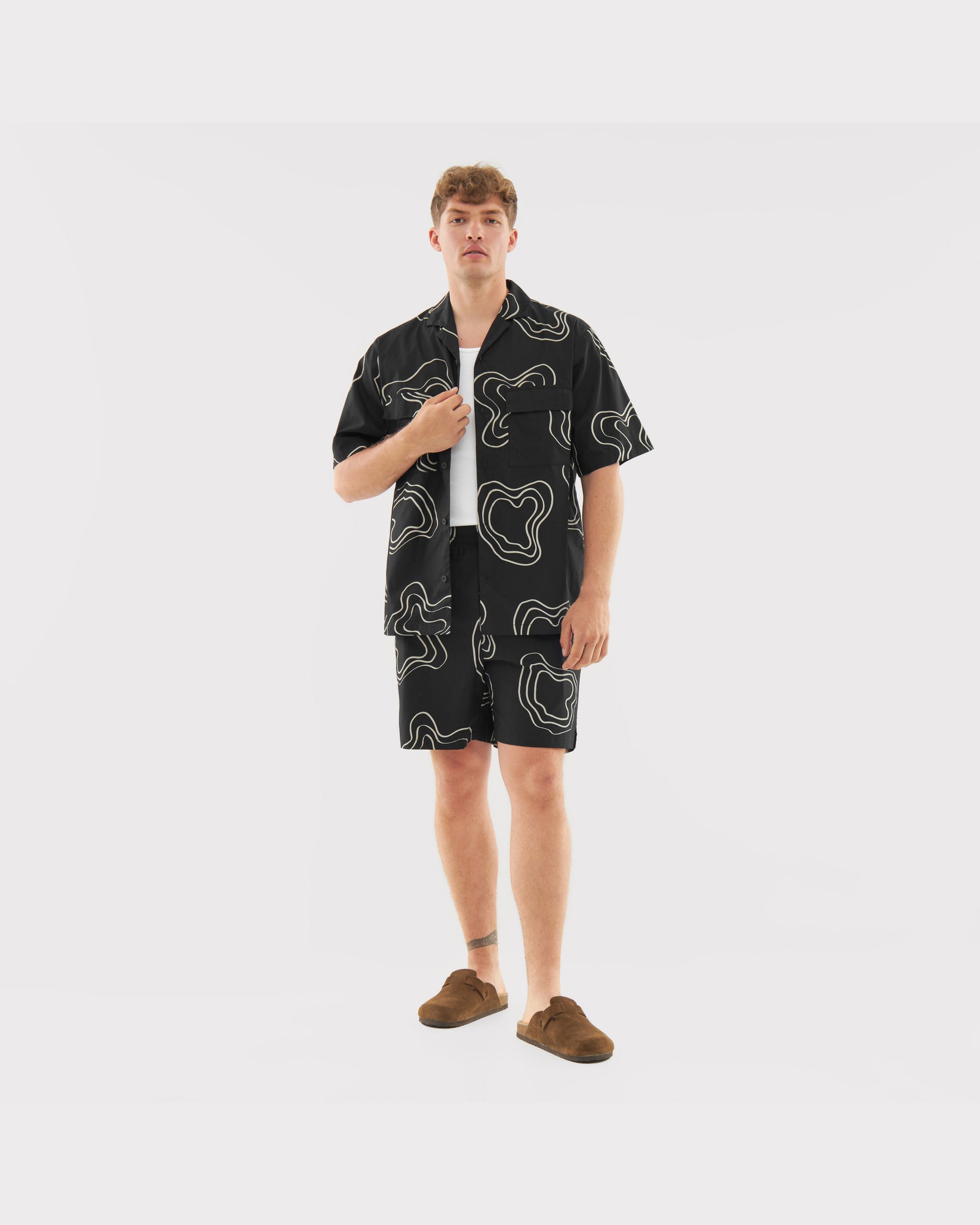 Mann trägt lockeres schwarzes Kurzarmhemd und Shorts mit weißem abstrakten Muster, weiße Unterziehshirt, braune Schuhe
