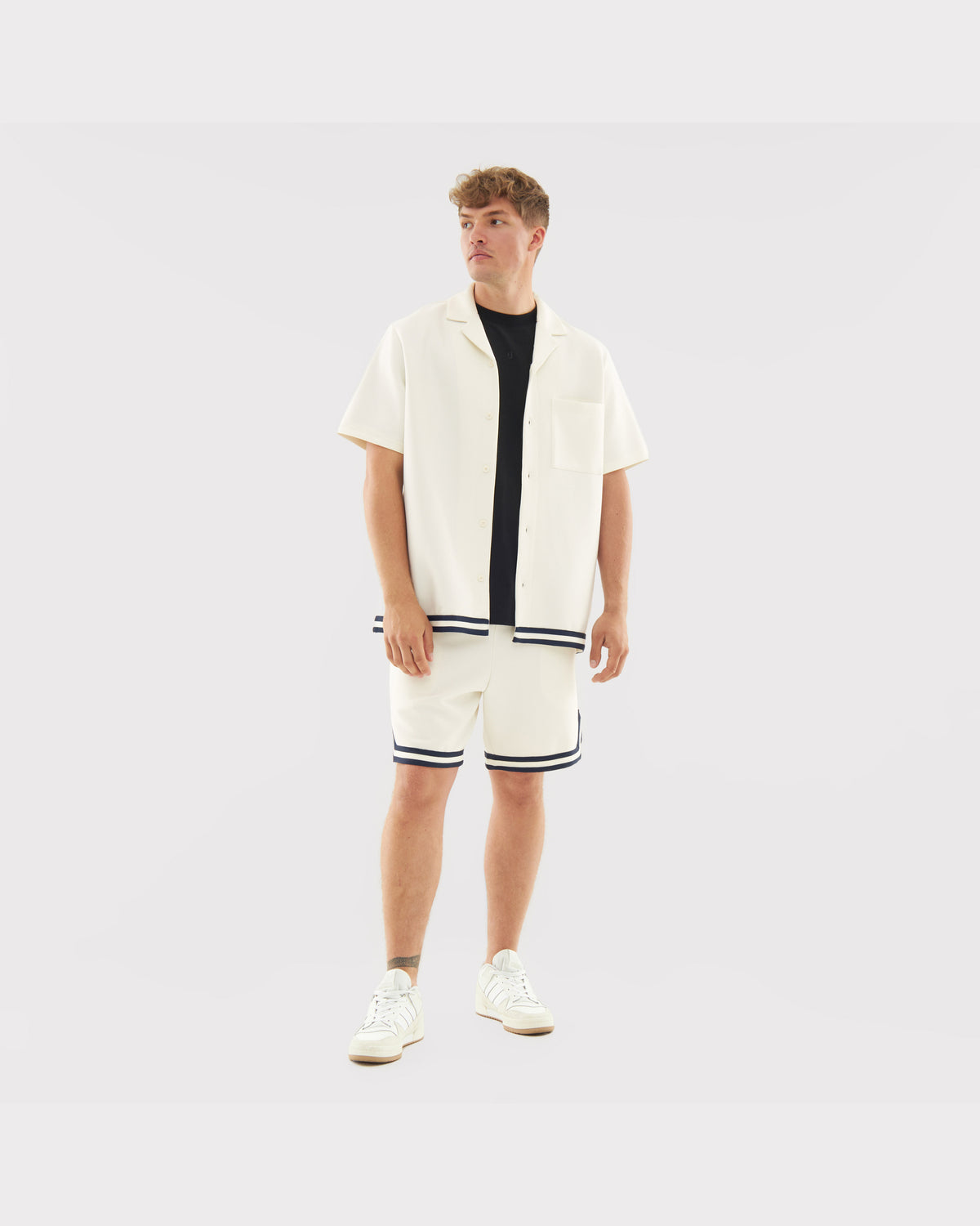 Mann trägt cremefarbenes Set 'Parker' mit kurzärmeligem Hemd und Shorts mit dunklen Streifen, weiße Sneakers