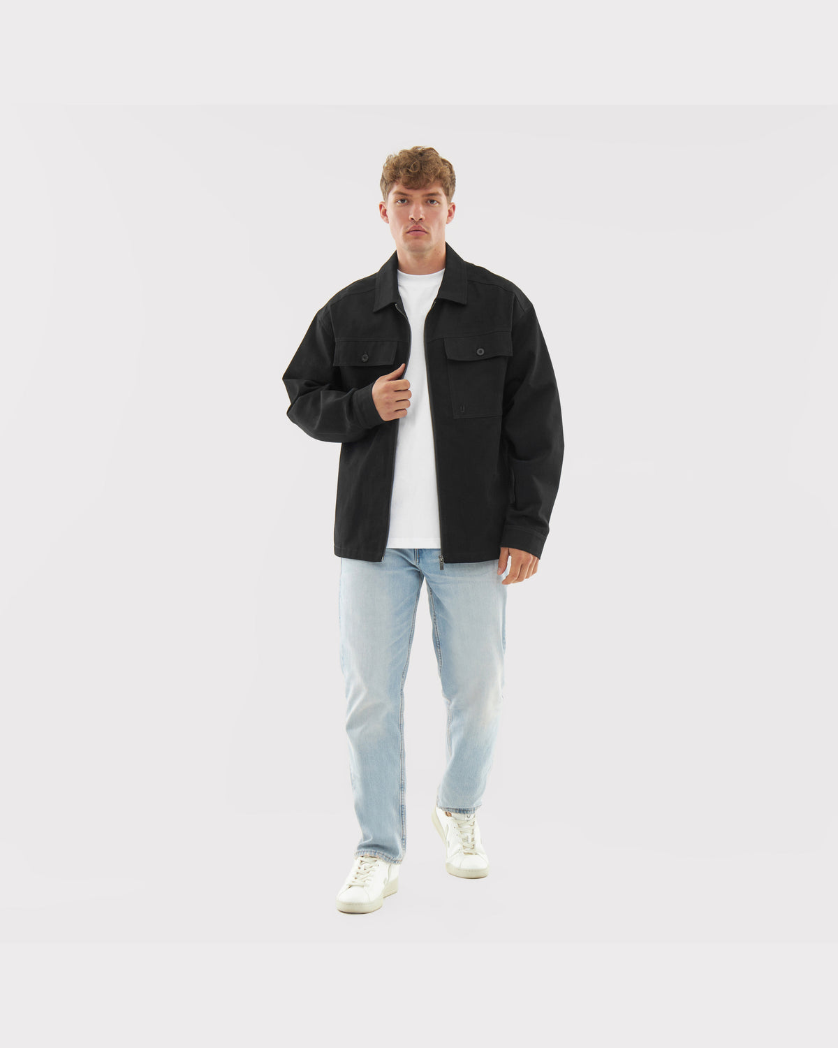 Mann trägt schwarze Baumwoll-Overshirt 'Cameron', weiße T-Shirt, helle Jeans und weiße Sneaker