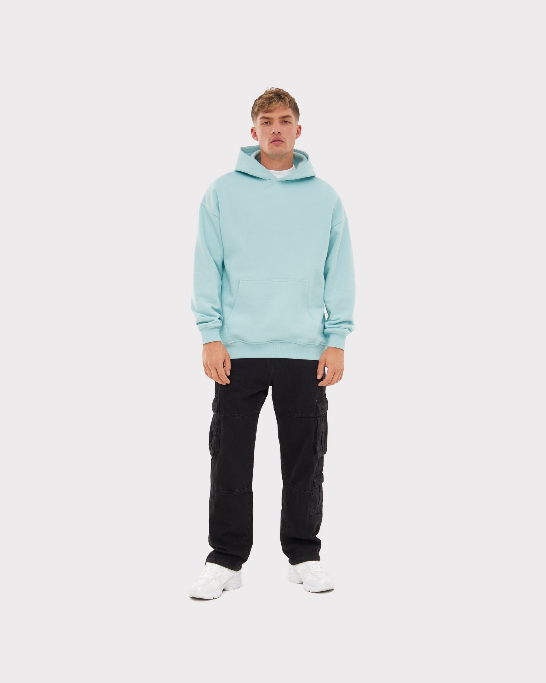 Mann trägt oversized Hoodie 'Cody' in eisblau mit schwarzer Cargo-Hose und weißen Sneakern