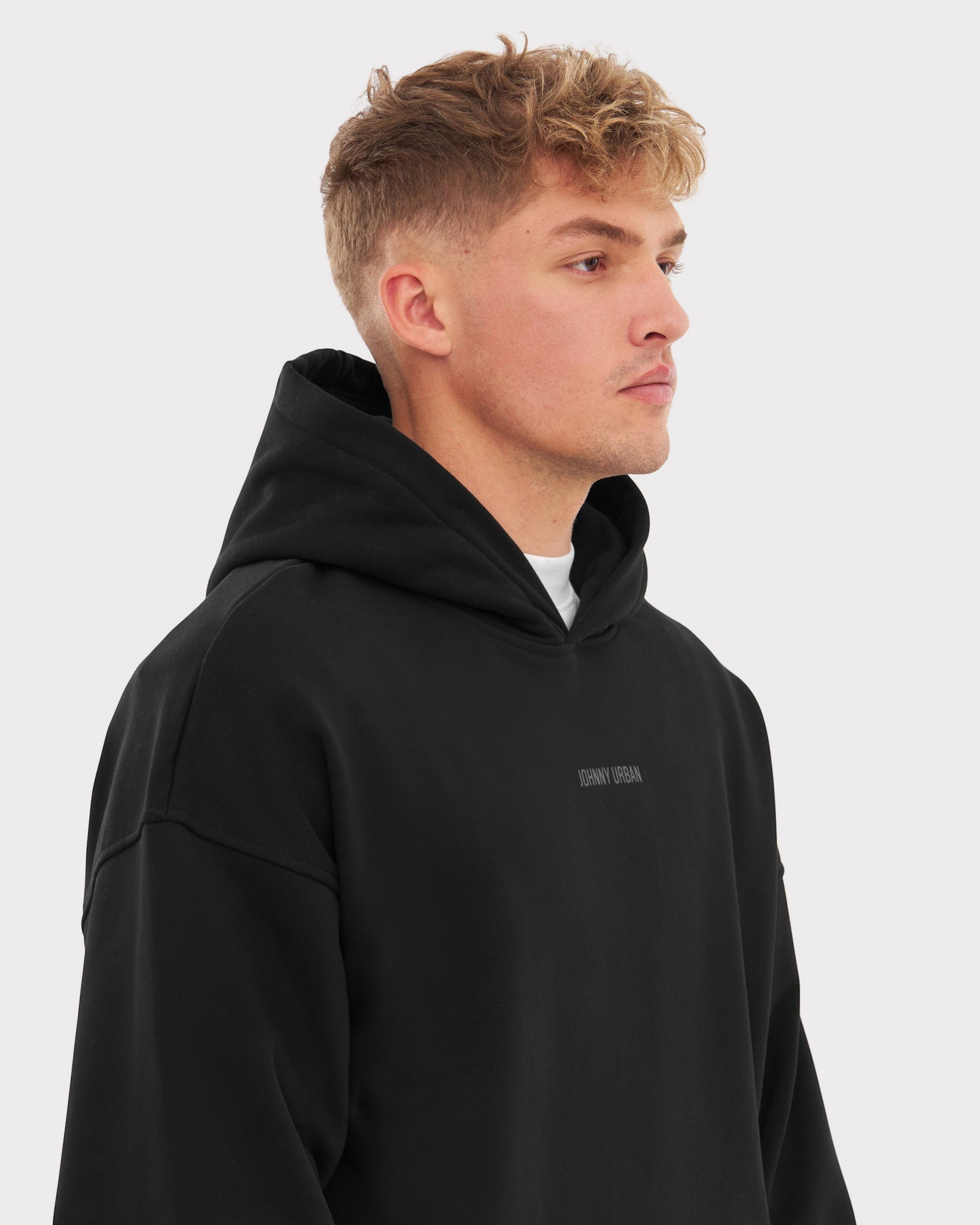 Junger Mann trägt schwarzen oversized Hoodie 'Cody' mit Kapuze und 'Johnny Urban' Print vorne