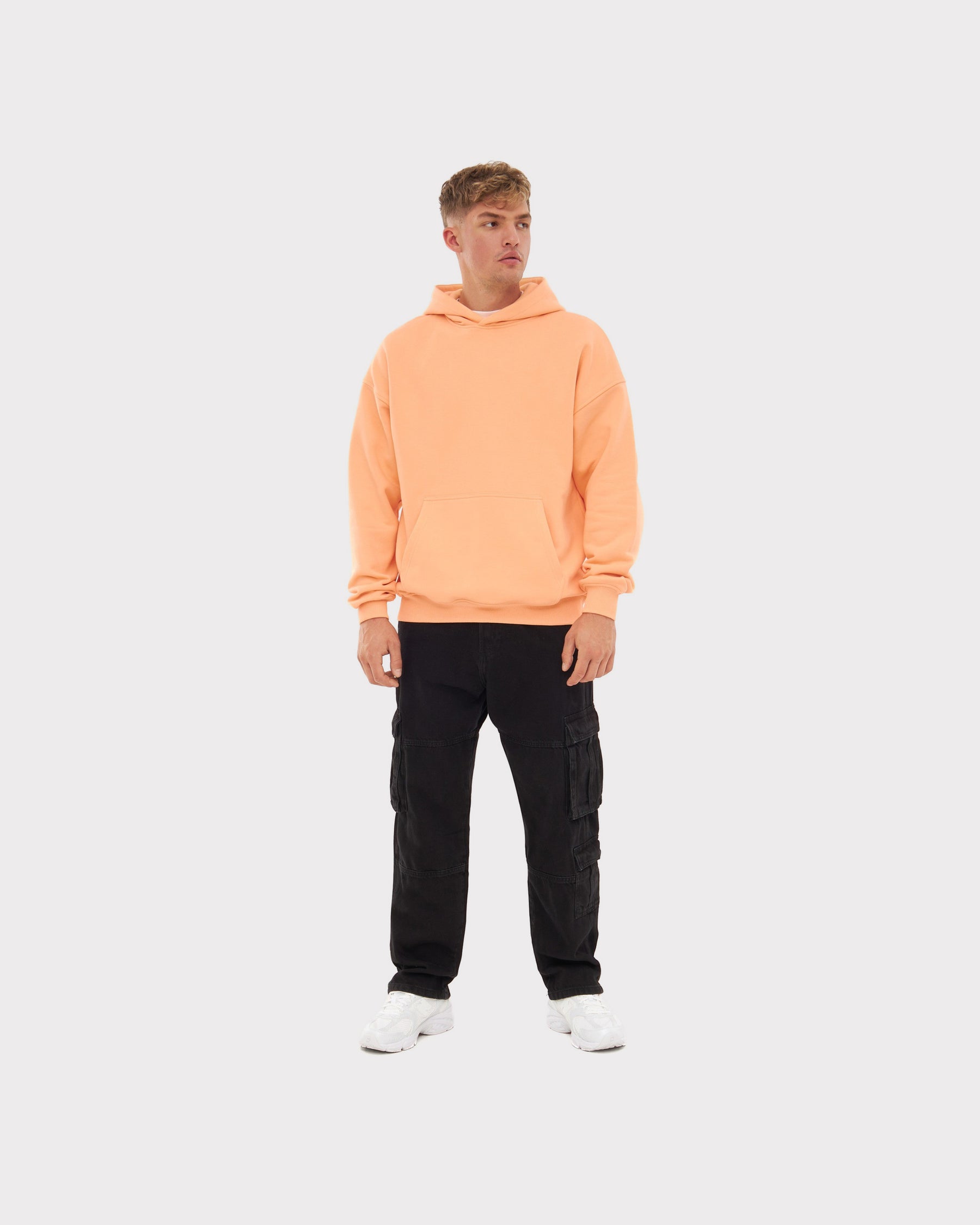 Mann mit oversized Hoodie 'Cody' in Pfirsich, schwarzen Cargo-Hosen und weißen Sneakers