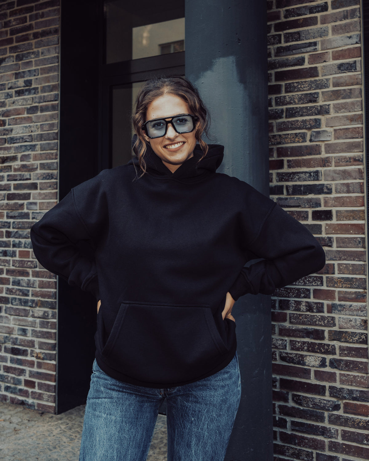 Frau trägt schwarzen Oversized Hoodie 'Cody' von Johnny Urban und blaue Jeans, steht vor Backsteinwand