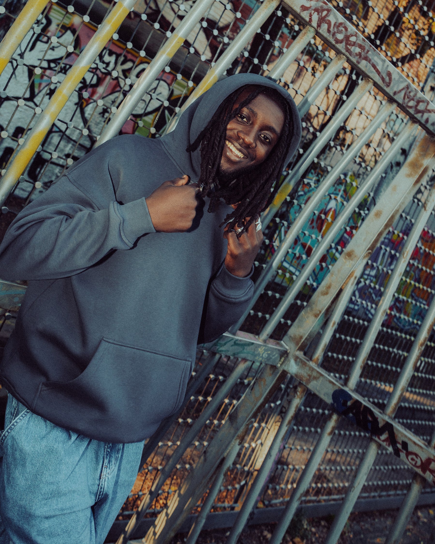 Mann trägt eine dunkelgraue oversized Hoodie 'Cody' von Johnny Urban vor einem mit Graffiti besprühten Metallzaun