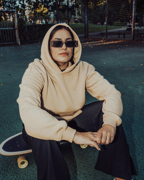 Frau trägt beigen Oversized-Hoodie 'Cody' und schwarze Sonnenbrille, sitzt auf Skateboard auf blauem Boden im Freien