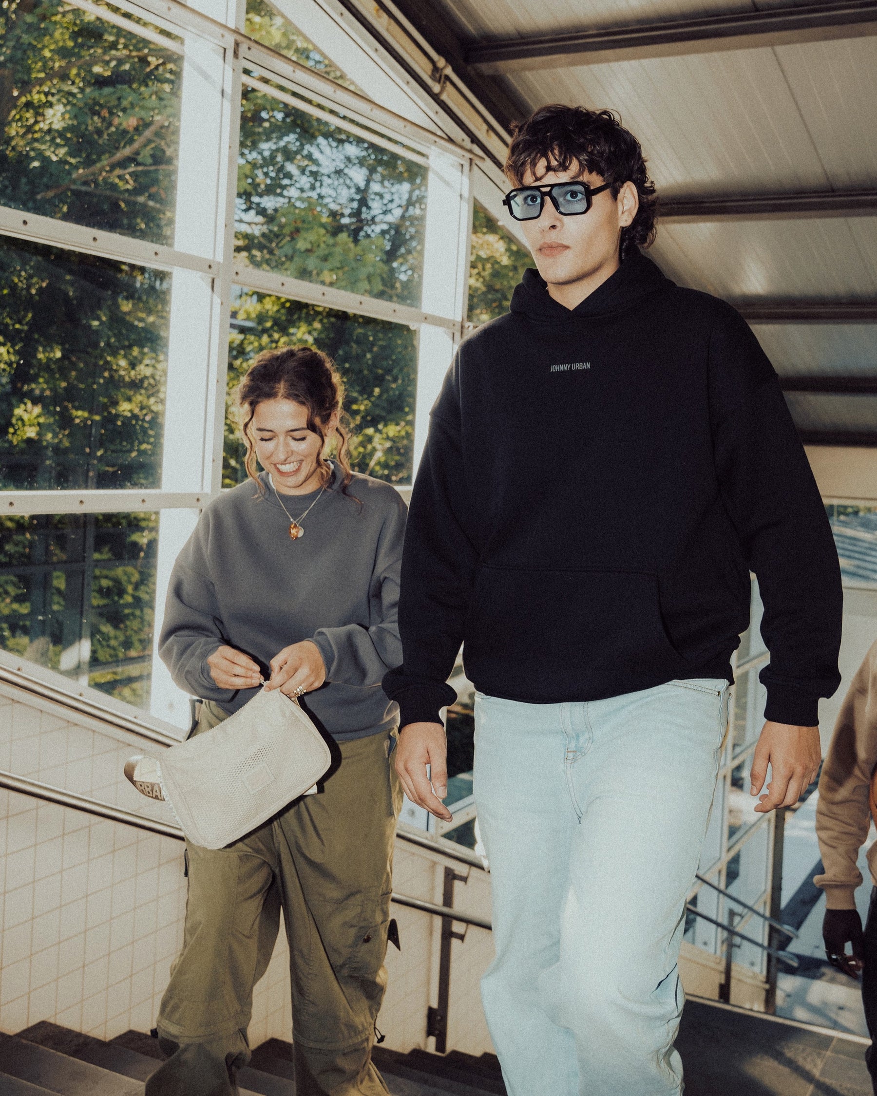 Junger Mann trägt schwarzen oversized Hoodie 'Cody' mit 'Johnny Urban' Print und helle Jeans, begleitet von zwei Personen auf Treppe