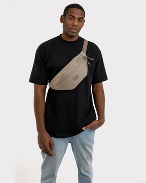 Mann trägt die große Bauchtasche ‚Erik Large‘ in desert grey über der Brust, dazu schwarzes T-Shirt und helle Jeans