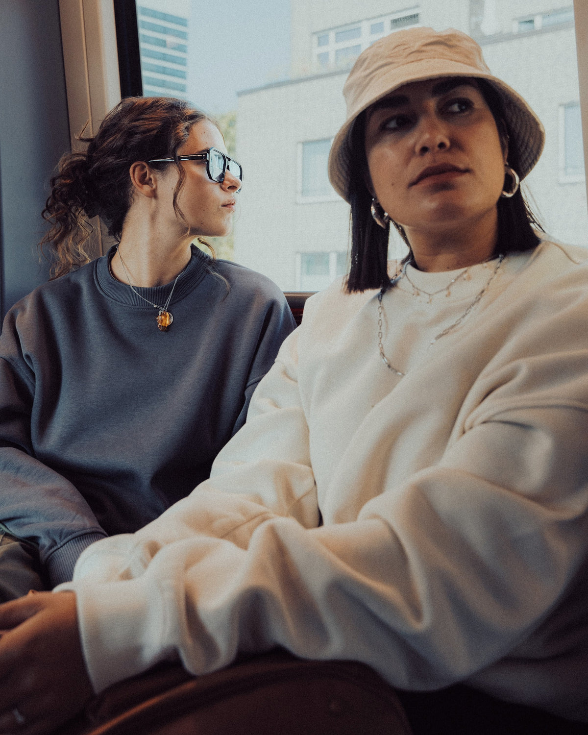 Zwei Frauen sitzen in der Straßenbahn, eine trägt ein graues Oversized Crewneck 'Carter', die andere einen weißen Pullover und einen Hut