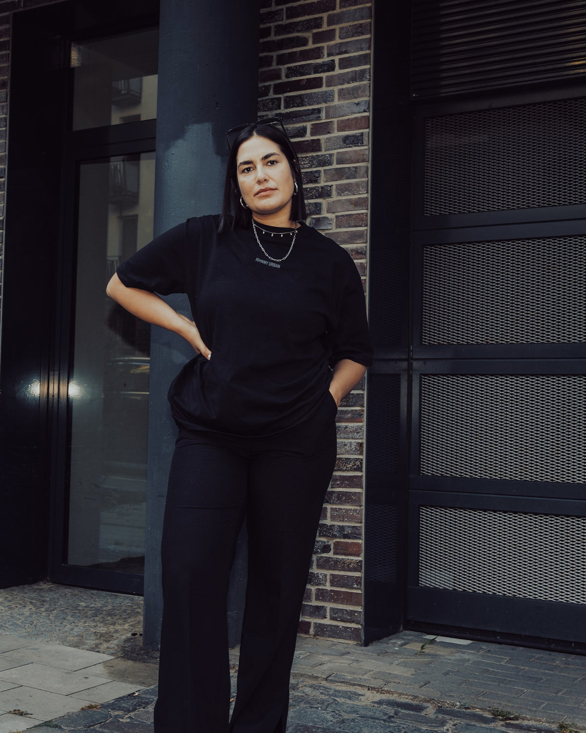 Frau trägt schwarz oversized T-Shirt 'Sammy Johnny Urban' und schwarze Hose vor urbanem Hintergrund