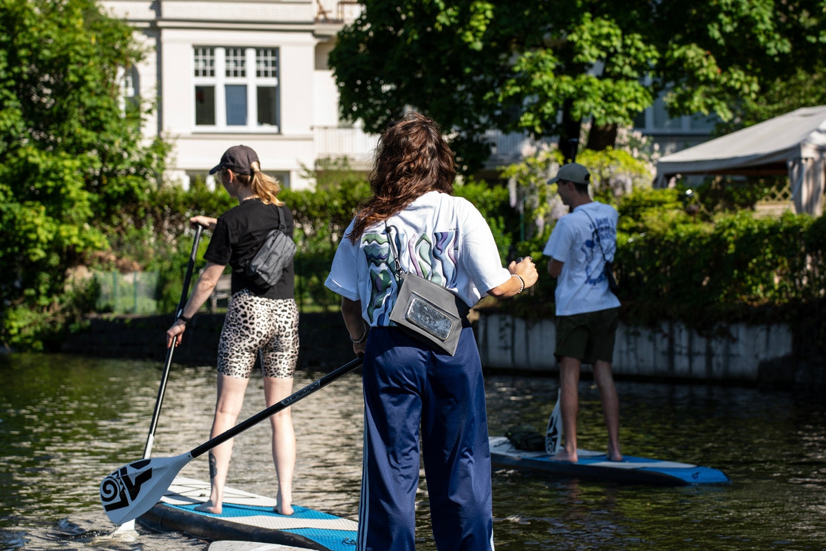 Frau steht auf einen Sup mit einem Paddel in der Hand und trägt eine wasserfeste Tasche.