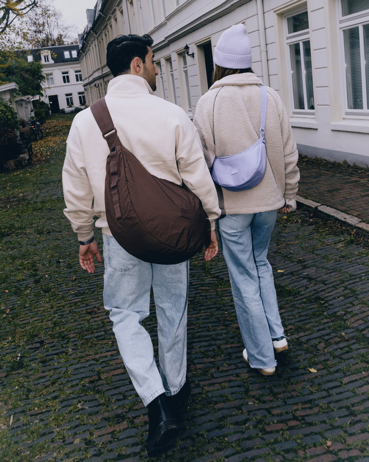 Zwei Personen gehen auf Kopfsteinpflaster, eine trägt die große braune Half Moon Bag 'Kaia XL', die andere eine kleine lila Half Moon Bag