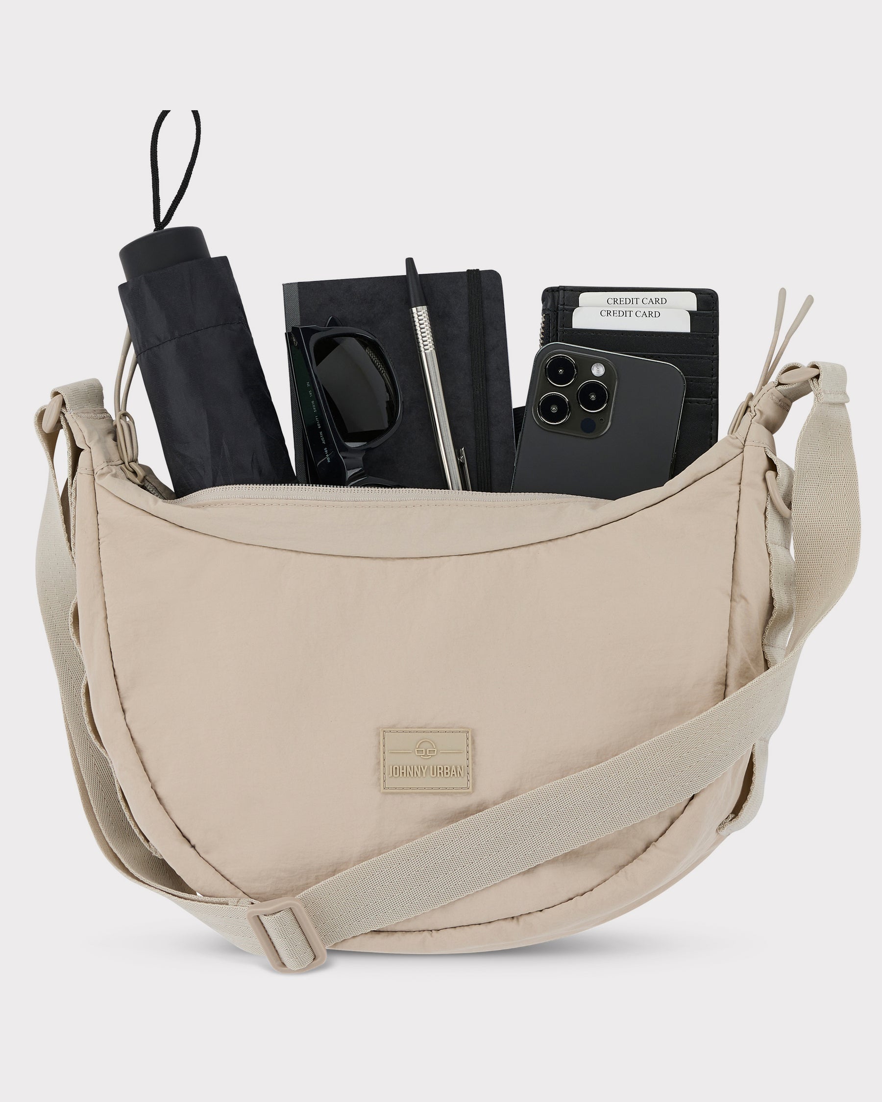 Halbmondtasche 'Kaia Medium' von Johnny Urban in Beige mit schwarzen Sonnenbrillen, Regenschirm, Notizbuch, Stift, Smartphone und Kreditkarten