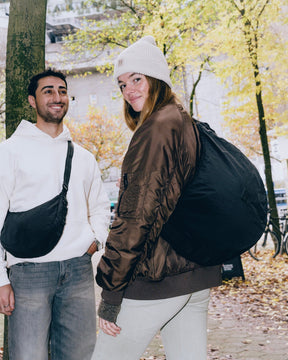 Mann mit hellbeigem Hoodie trägt schwarze Half Moon Bag Kaia XL crossbody, Frau mit brauner Jacke und weißer Mütze trägt sie als Rucksack