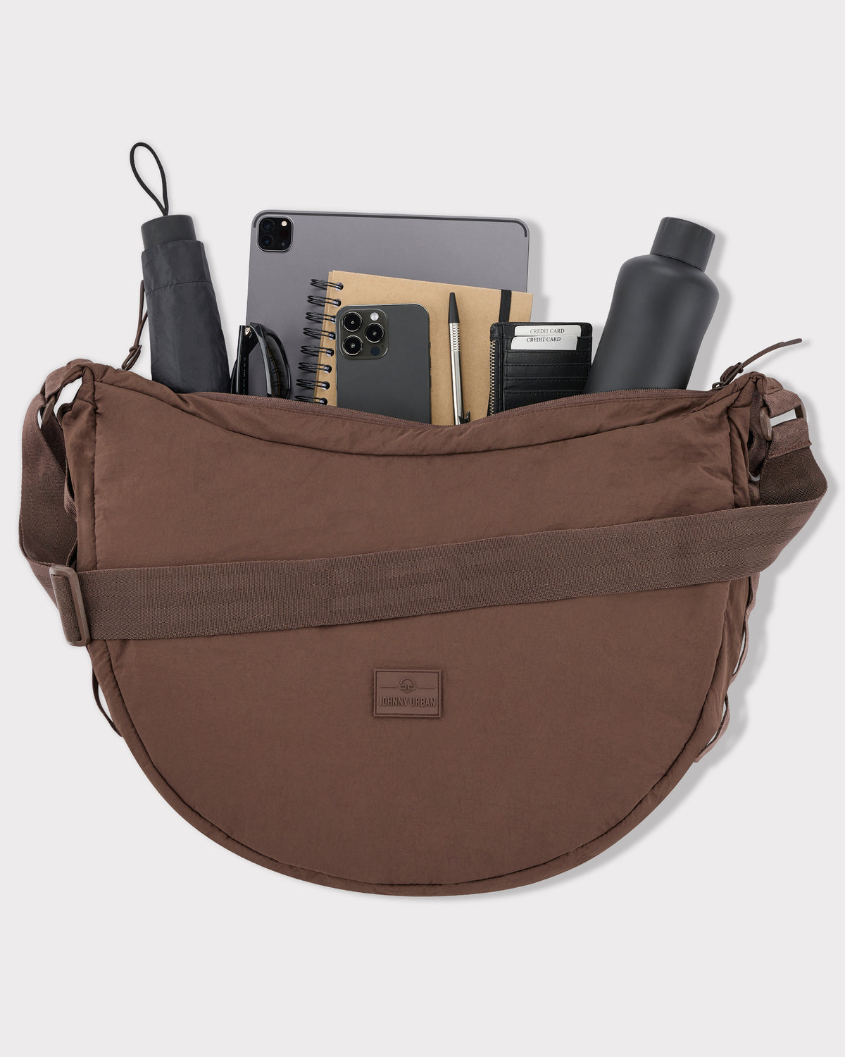 Halbmondtasche Kaia XL von Johnny Urban in Braun mit Laptop, Smartphone, Notizbuch, Stift, Kreditkartenetui, Wasserflasche, Regenschirm und Sonnenbrille gefüllt