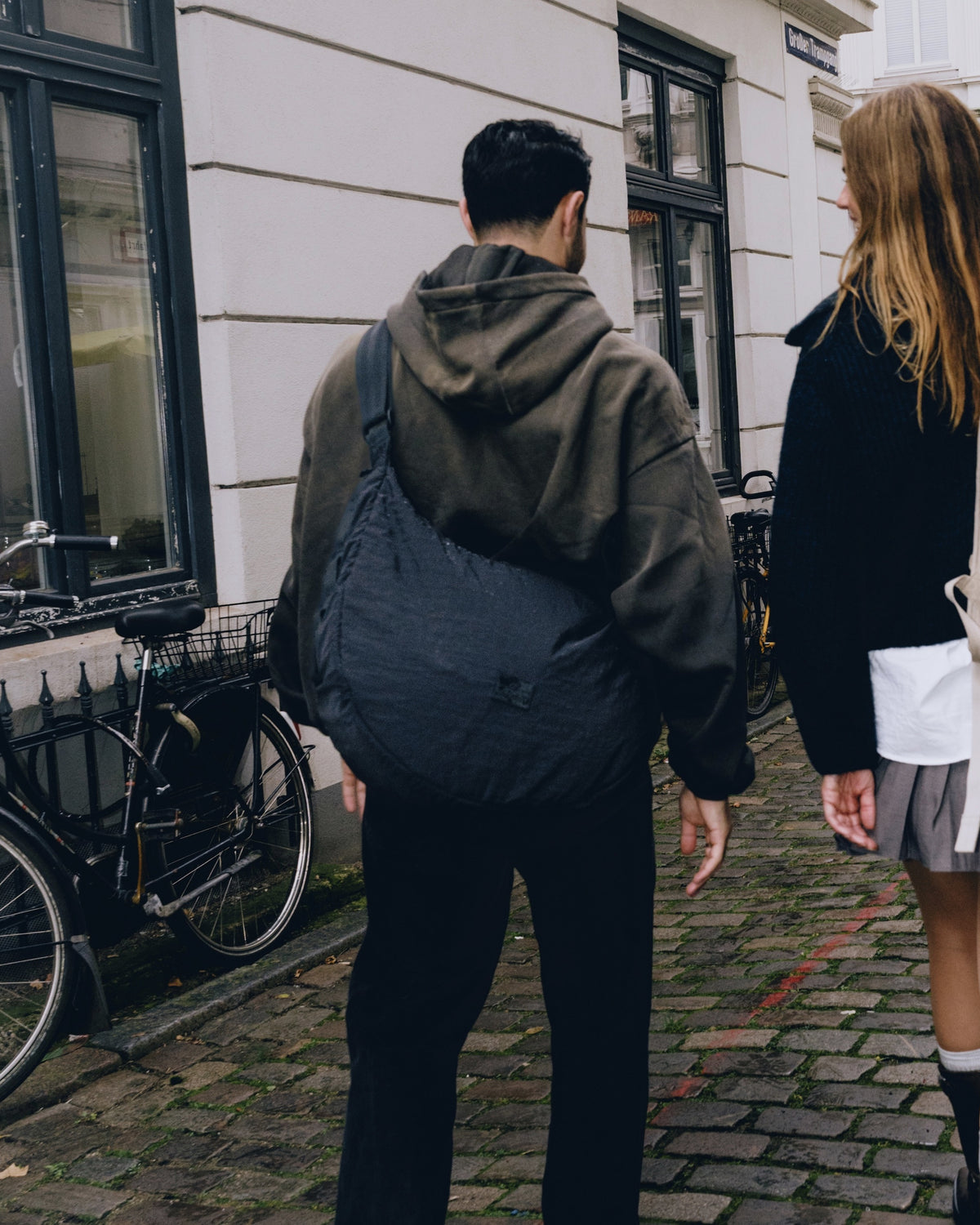Mann trägt schwarze Half Moon Bag 'Kaia XL' von Johnny Urban über der Schulter, geht mit Frau auf Kopfsteinpflasterstraße