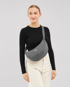 Frau mit schwarzem Langarmshirt und hellen Hosen trägt halbrunde, graue Half Moon Bag 'Ally Medium' quer über dem Oberkörper