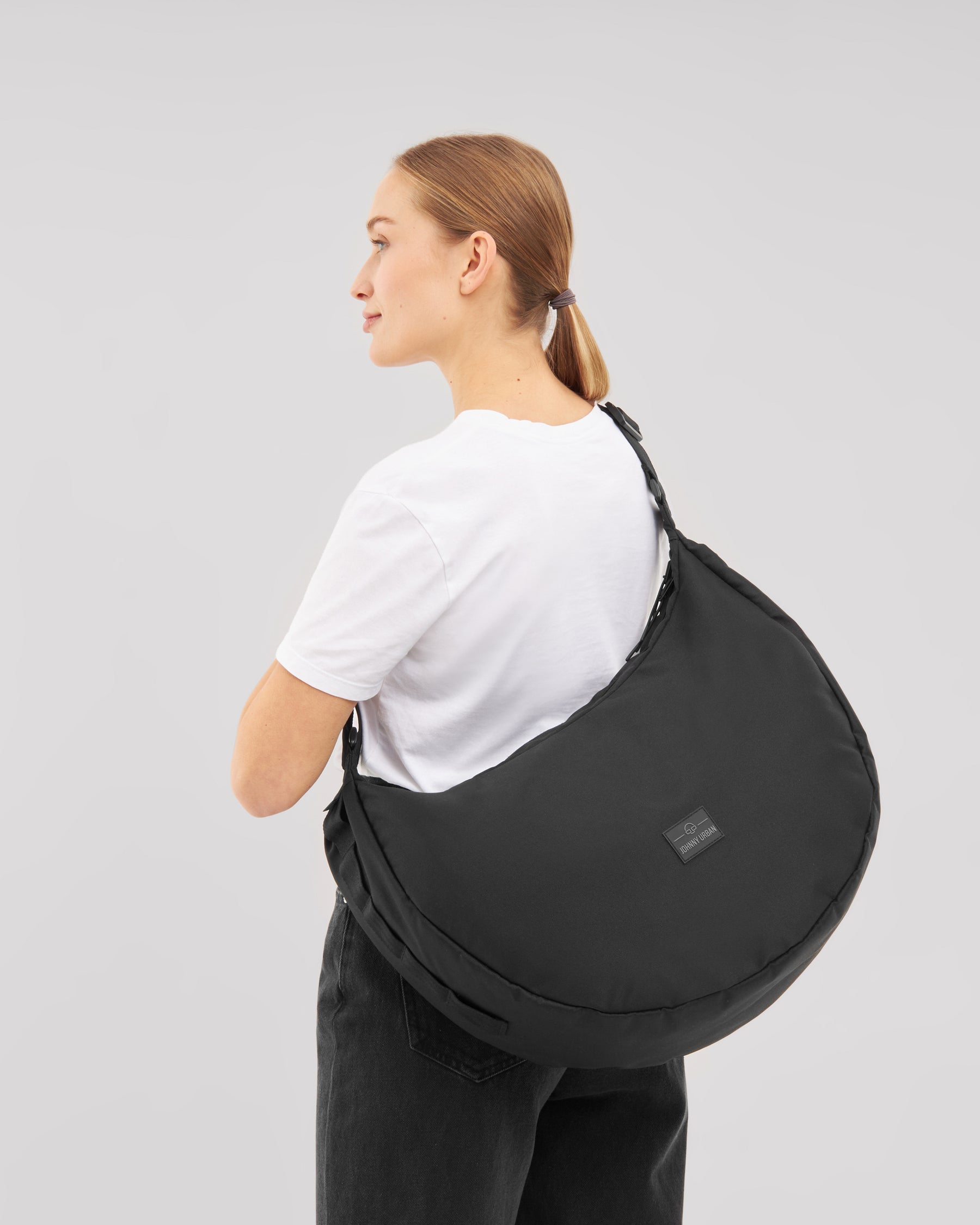 Frau trägt schwarze Half Moon Bag 'Ally XL' Schultertasche von Johnny Urban über weißem T-Shirt und schwarzer Hose