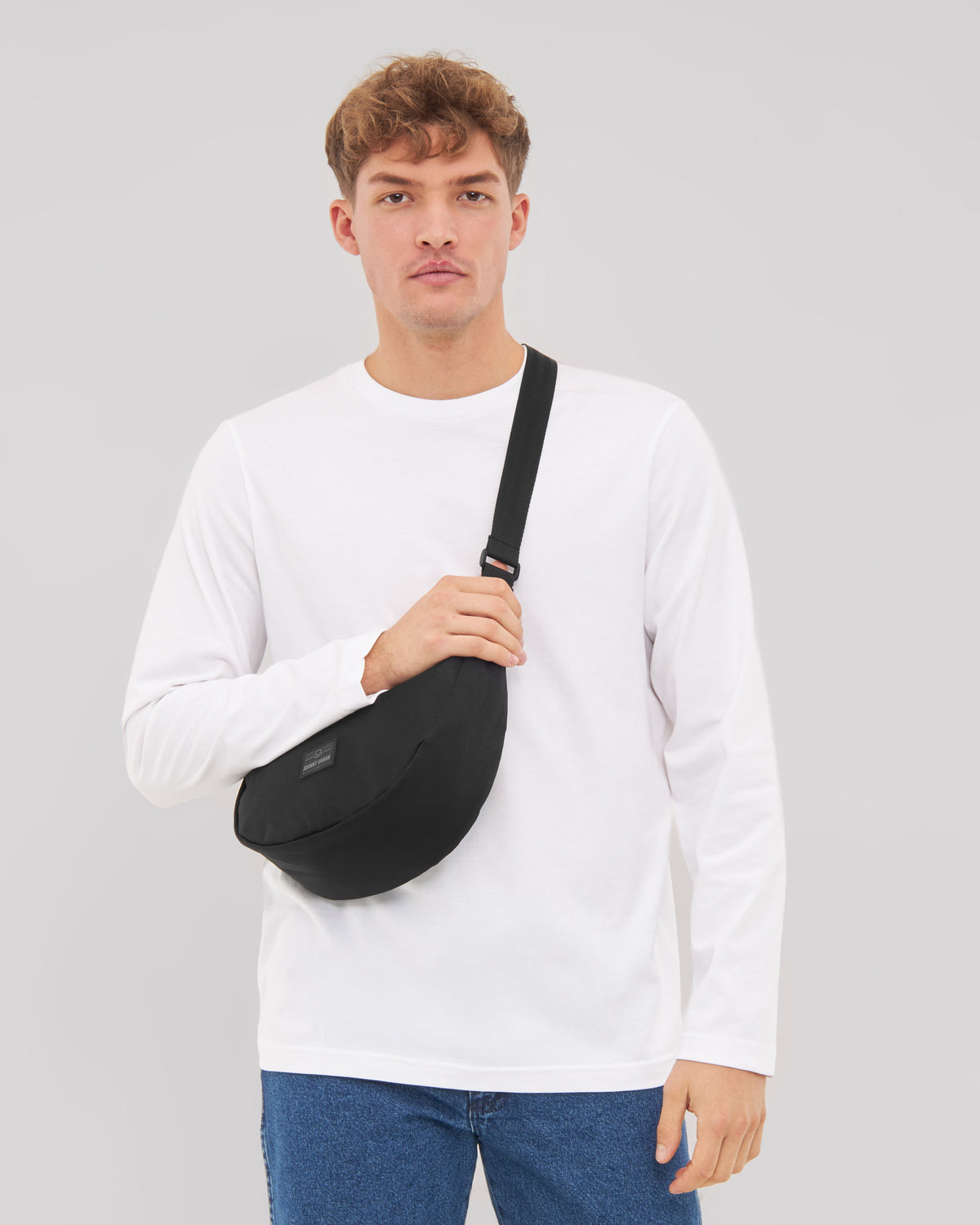 Mann mit weißem Langarmshirt und blauen Jeans trägt schwarze Half Moon Bag 'Ally Medium' quer am Oberkörper