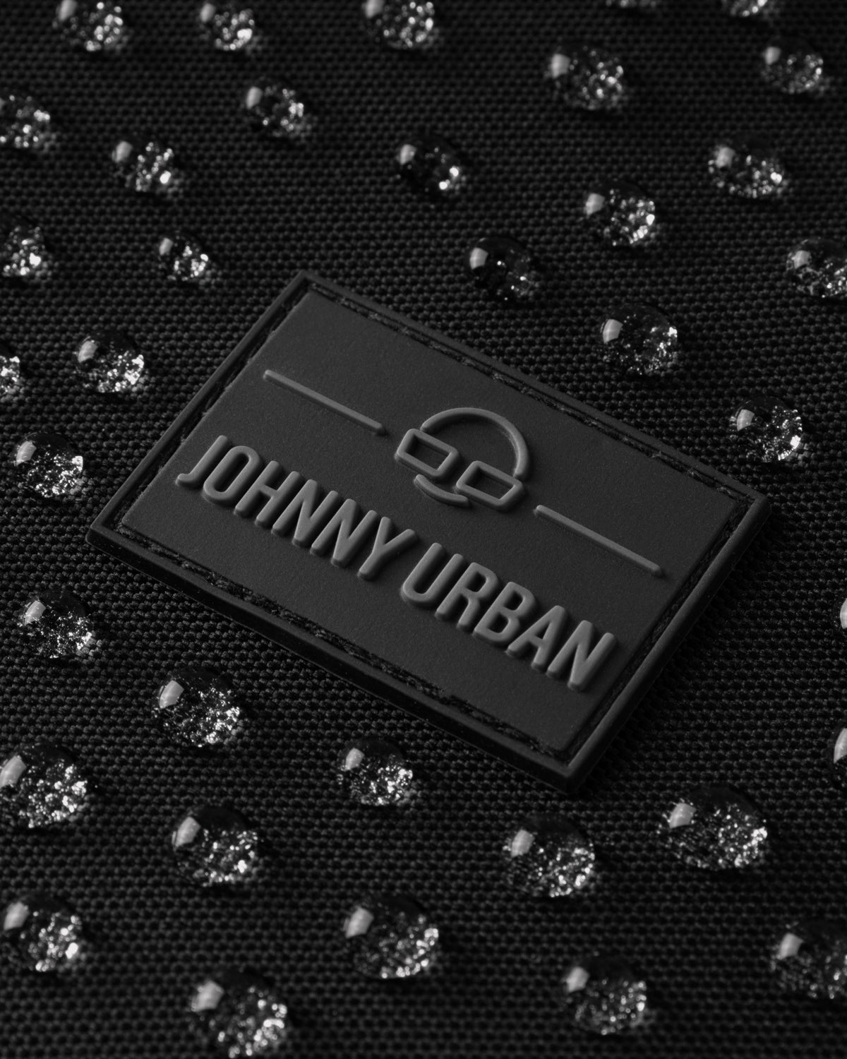 Nahe Ansicht eines schwarzen Johnny Urban Labels auf wasserabweisendem Material mit Wassertropfen auf der Halbmondtasche Ally XL
