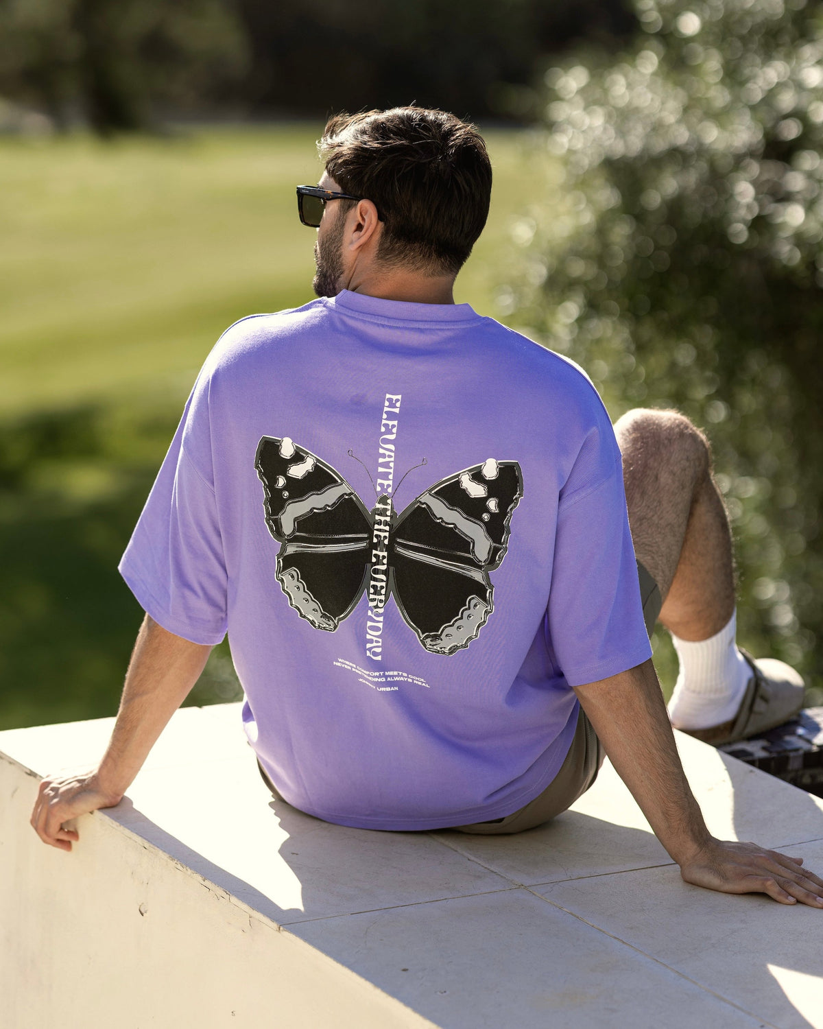 Mann sitzt draußen, trägt lila Oversize-T-Shirt 'Sammy Chrome Butterfly' mit großem schwarzem Schmetterlingsdruck auf dem Rücken