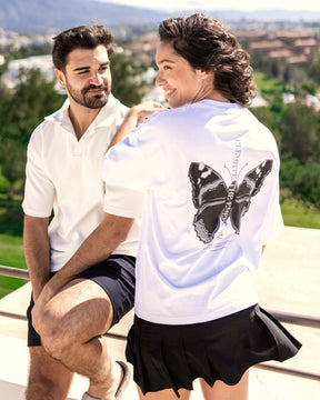 Frau trägt weißes Oversize-T-Shirt 'Sammy Chrome Butterfly' mit schwarzem Schmetterlingsmotiv auf dem Rücken, Mann daneben in weißem Poloshirt und schwarzen Shorts