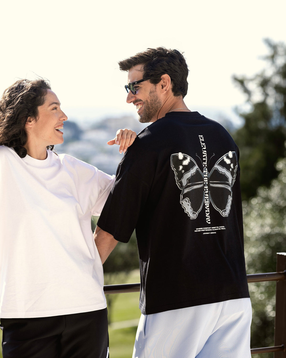 Mann trägt schwarzes Oversize-T-Shirt 'Sammy Chrome Butterfly' mit großem Schmetterlingsprint auf dem Rücken, Frau daneben lächelnd
