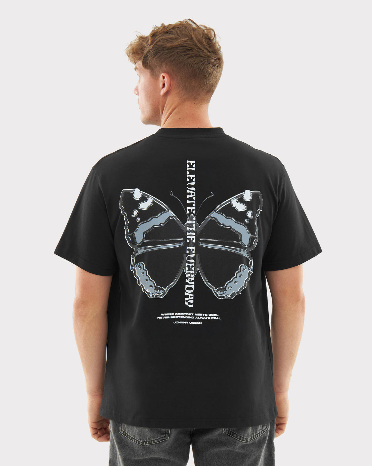 Mann trägt schwarzen Oversize-T-Shirt 'Sammy Chrome Butterfly' mit grauem Schmetterlingsprint und senkrechtem Schriftzug 'ELEVATE THE EVERY DAY' auf dem Rücken