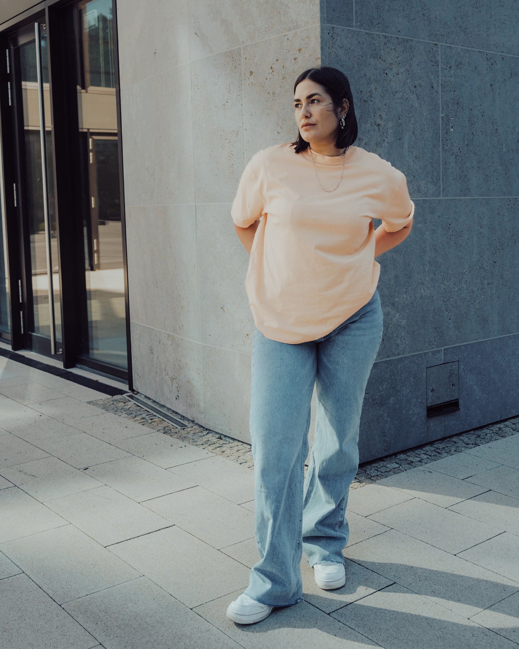 Frau trägt Oversize-T-Shirt 'Sammy' in Pfirsich-Orange und weite blaue Jeans vor moderner Fassade