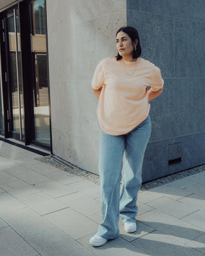 Frau trägt Oversize-T-Shirt 'Sammy' in Pfirsich-Orange und weite blaue Jeans vor moderner Fassade