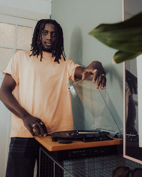 Mann mit Dreadlocks in peachfarbenem Oversize-T-Shirt 'Sammy' von Johnny Urban neben Plattenspieler