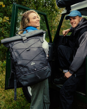 Frau mit dunkelgrauem Reiserucksack 'Allen Large Travel' von Johnny Urban, Mann sitzt in grünem Geländewagen