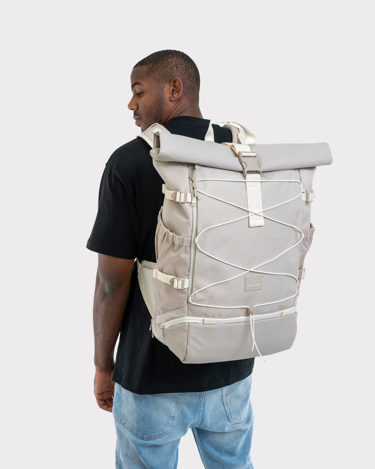 Mann mit schwarzem T-Shirt und Jeans trägt beigefarbenen Johnny Urban Reiserucksack 'Allen Large Travel' mit gerolltem Verschluss und elastischen Schnüren