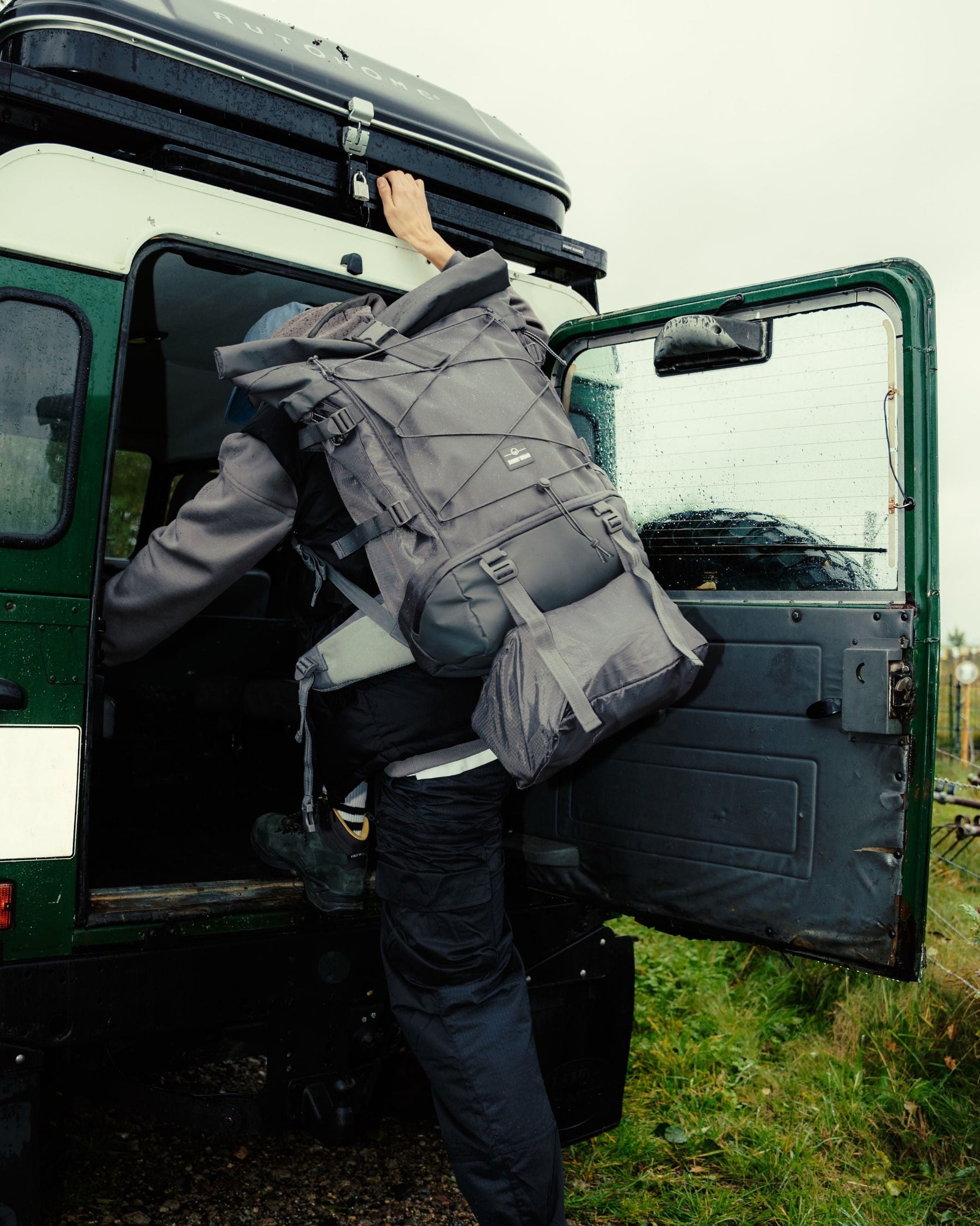 Person klettert mit großem grauem Johnny Urban Allen XL Travel Reiserucksack in grünes Geländefahrzeug